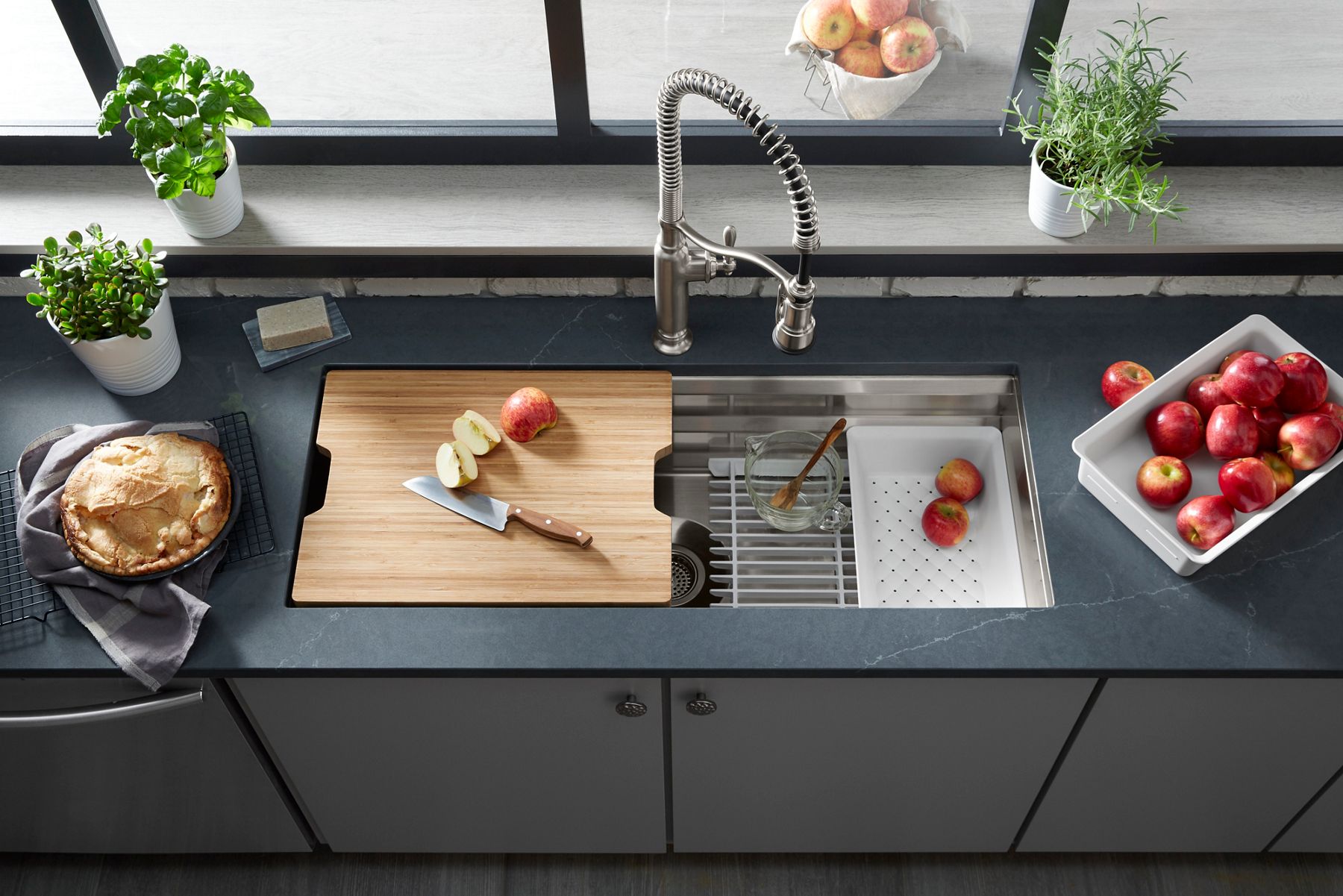 Overhead view of a workstation sink with a cutting board with a knife and apple slices on the left and a colander and more apple slices on the right.