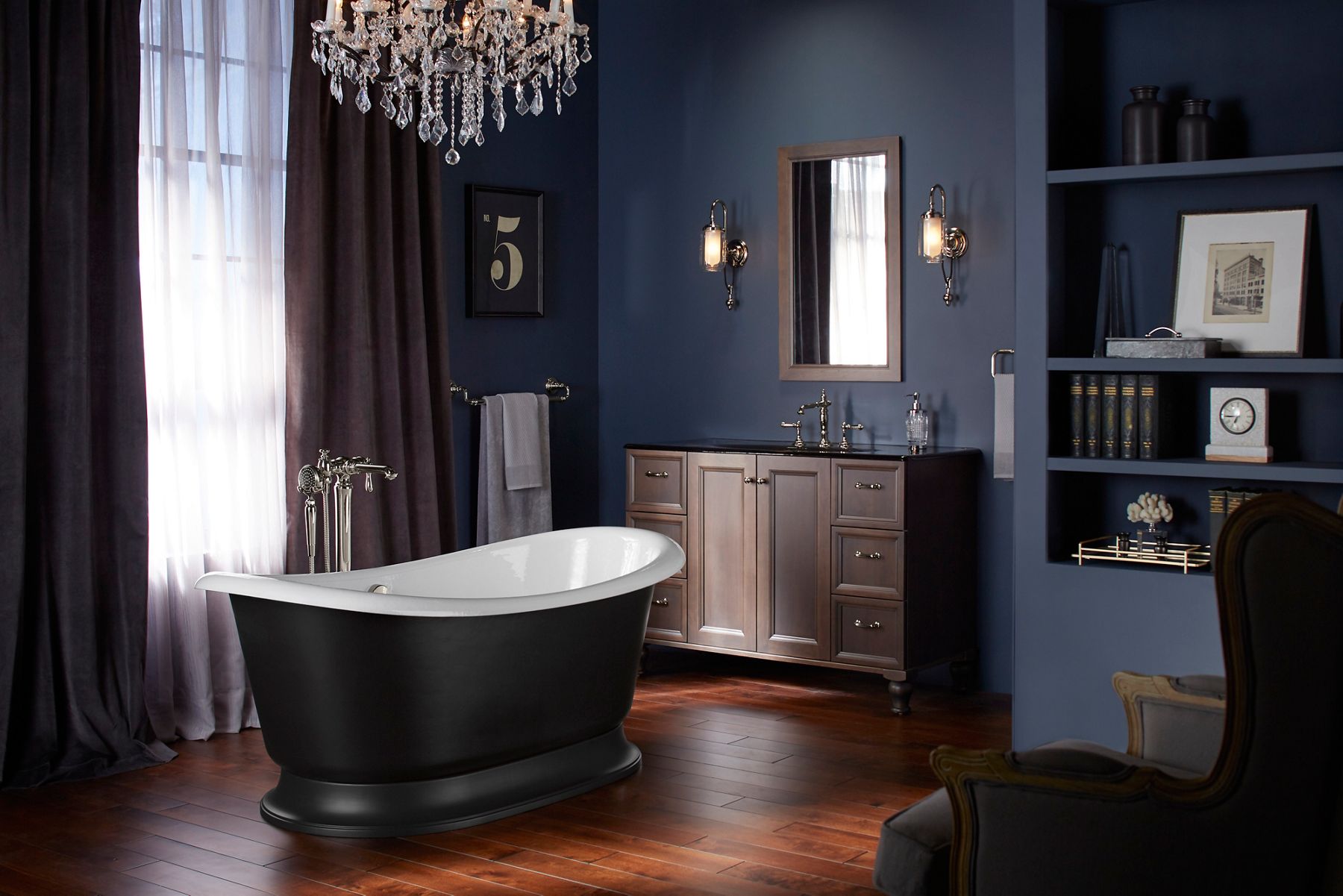 A freestanding bath with a black exterior and white interior in a darkened room with a wood vanity and chandelier, and light coming in from a nearby window.