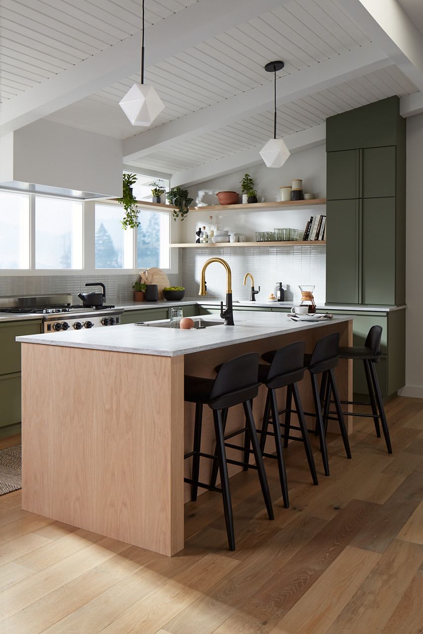 Wide image of a sunny kitchen with earthy green cabinet colors and natural wood. The large island features a centered sink with two-tone Kohler faucet in the center, across from a wall of windows and open storage shelves.