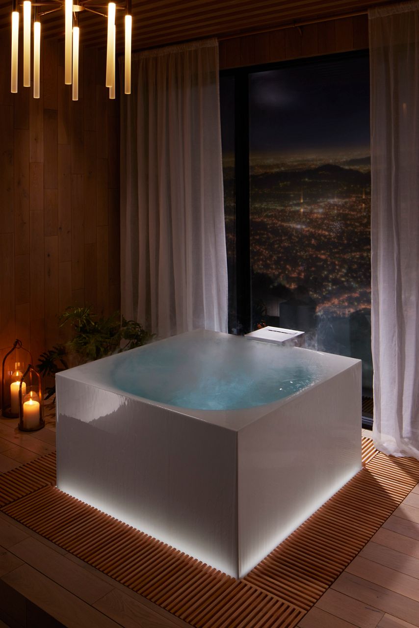 Three-quarter overhead angle image of a white square-shaped KOHLER Stillness bath filled perfectly filled with water, illuminated with light inside and around the edges, with steam rising up from the water. The bath is installed in a room with large windows overlooking a city.