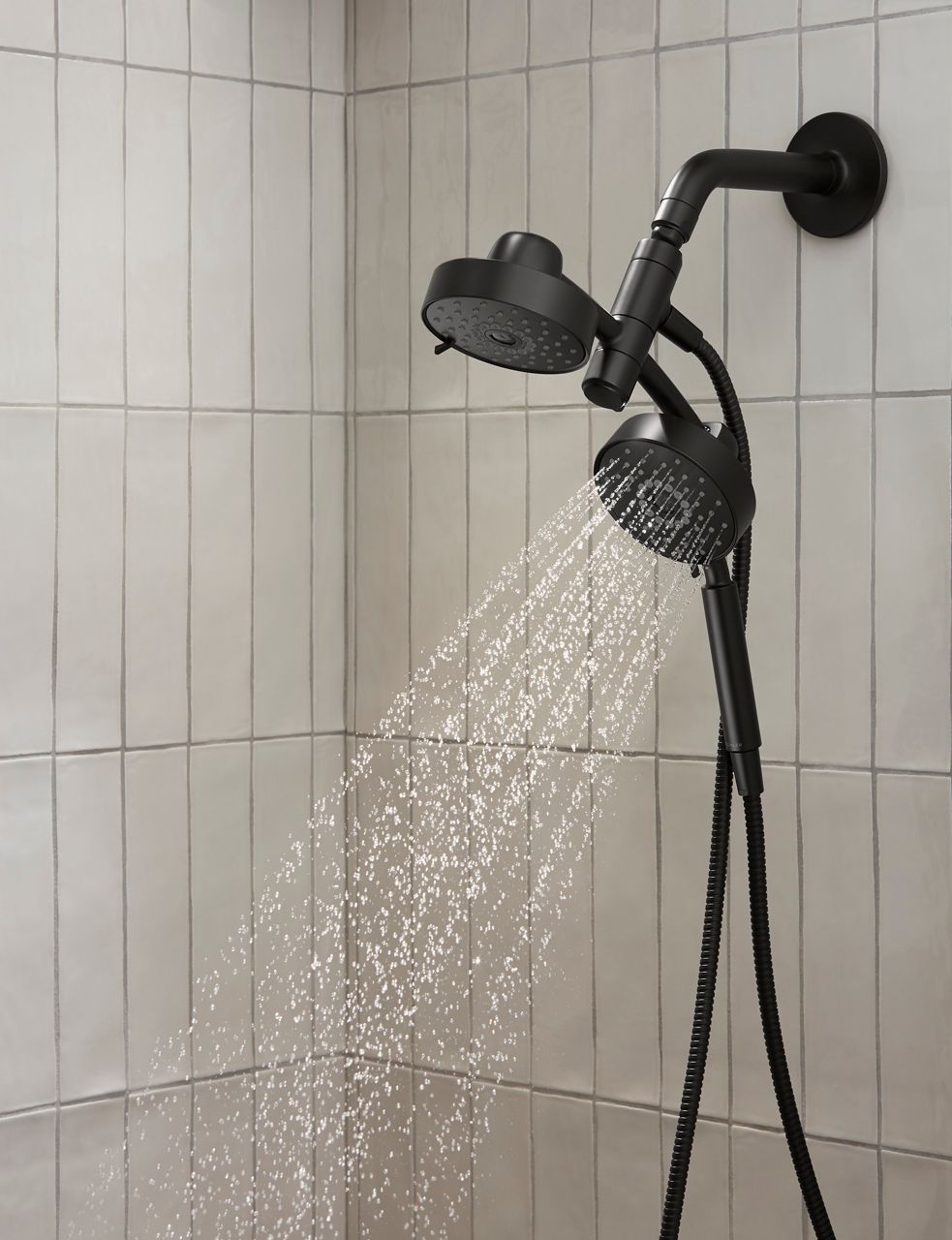 Black showerhead and hand shower with spray mounted on a white tile wall.