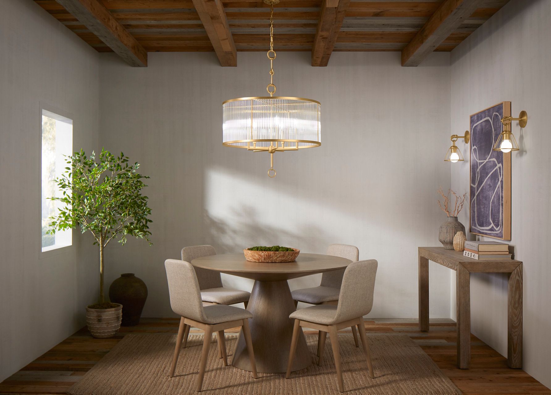 Wide image of a full dining room space, featuring a large circular Kohler chandelier of glass and vibrant brass above a modern circular wooden dining room table and four chairs. Matching brass wall sconces are above a wood buffet table again the wall opposite a large window, allowing sunlight to fill the room.