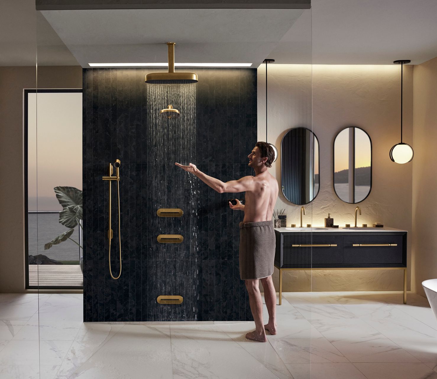 Image of a man standing next to his KOHLER Statement and Anthem shower system with the rainhead shower turned on and he's reaching out to feel the water coming down. He's in a towel and about to enter the shower. 