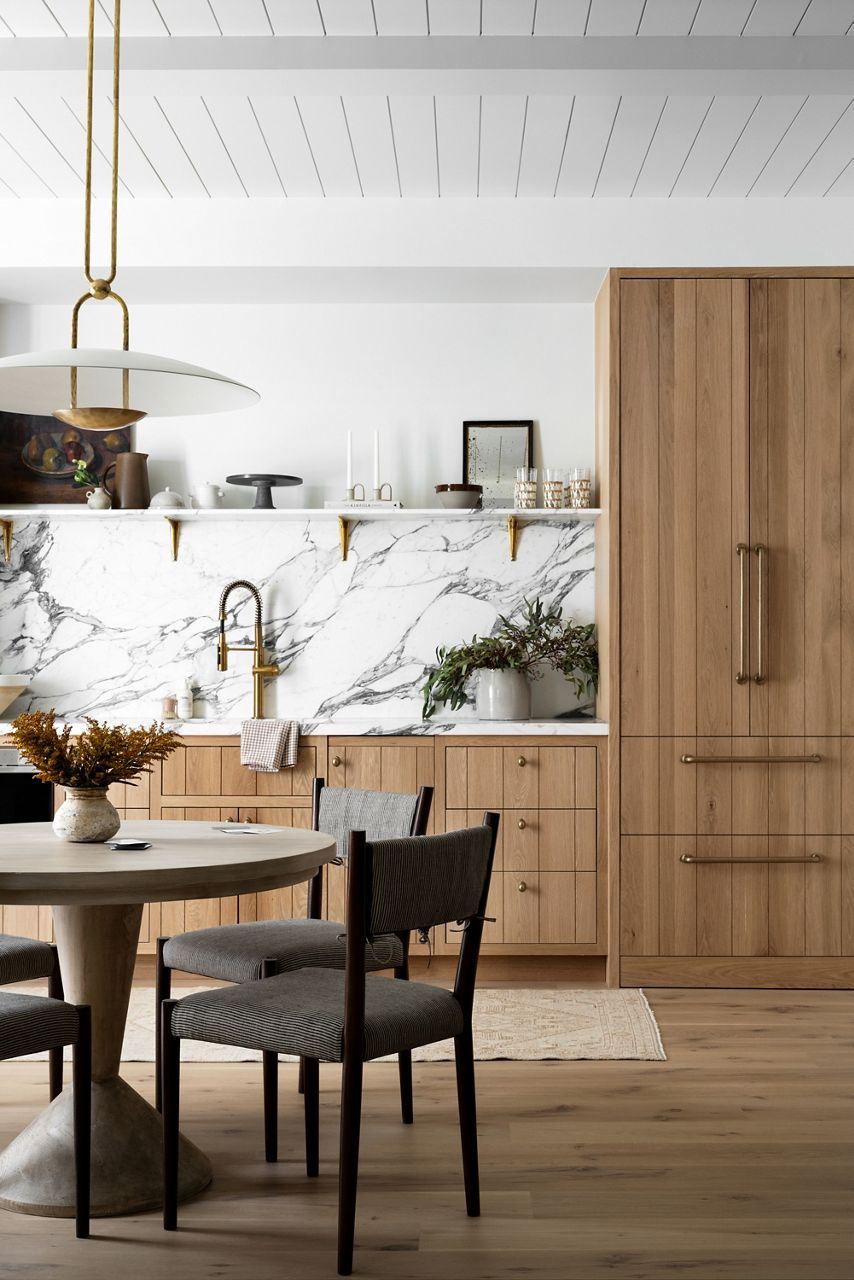 Image of a white marble and natural wood kitchenette design with dining table in the foreground and a row of kitchen cabinets and kitchen sink on the back wall of the room.