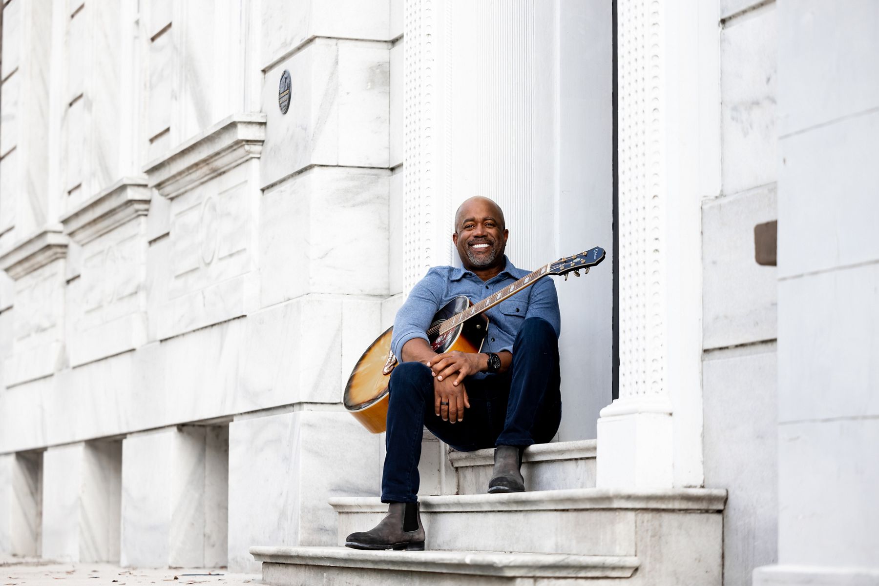Portrait of Darius Rucker, musician and tv host, sitting on the stone-carved stairs of a building, holding a guitar on his lap.