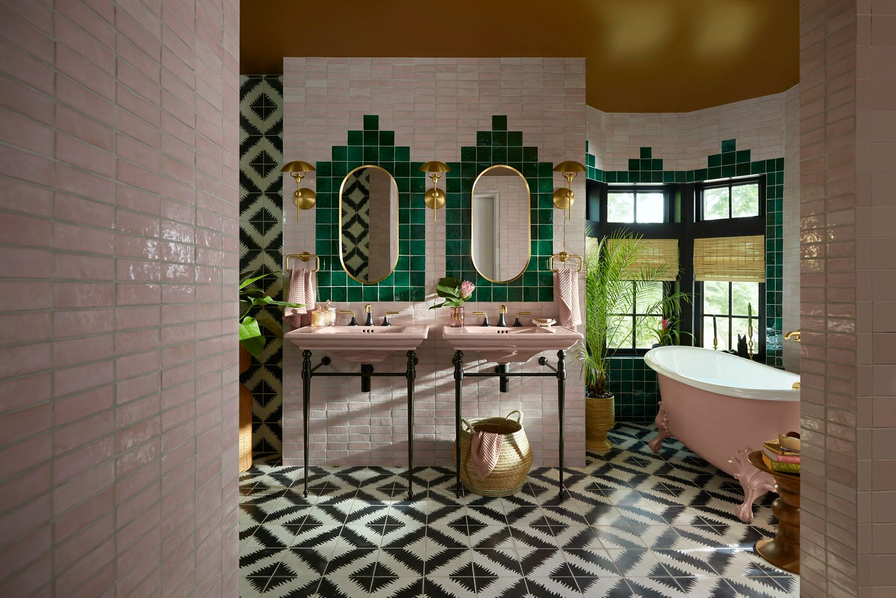 A bathroom designed by Justina Blakeney using two console sinks, a freestanding cast iron bath, and wall tile in a peachblow color, along with tile in an emerald green and black accents.
