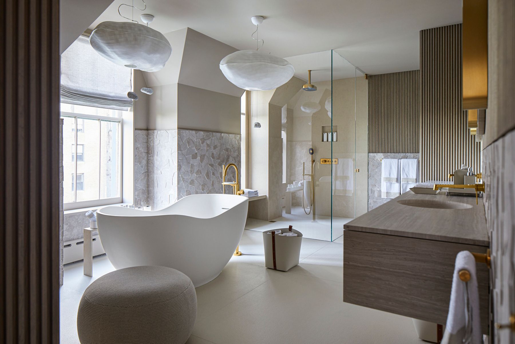  A large sun-filled bathroom in neutral tones with gold accents with a freestanding bath on the left, a glass-walled shower behind it, and a double vanity on the right.