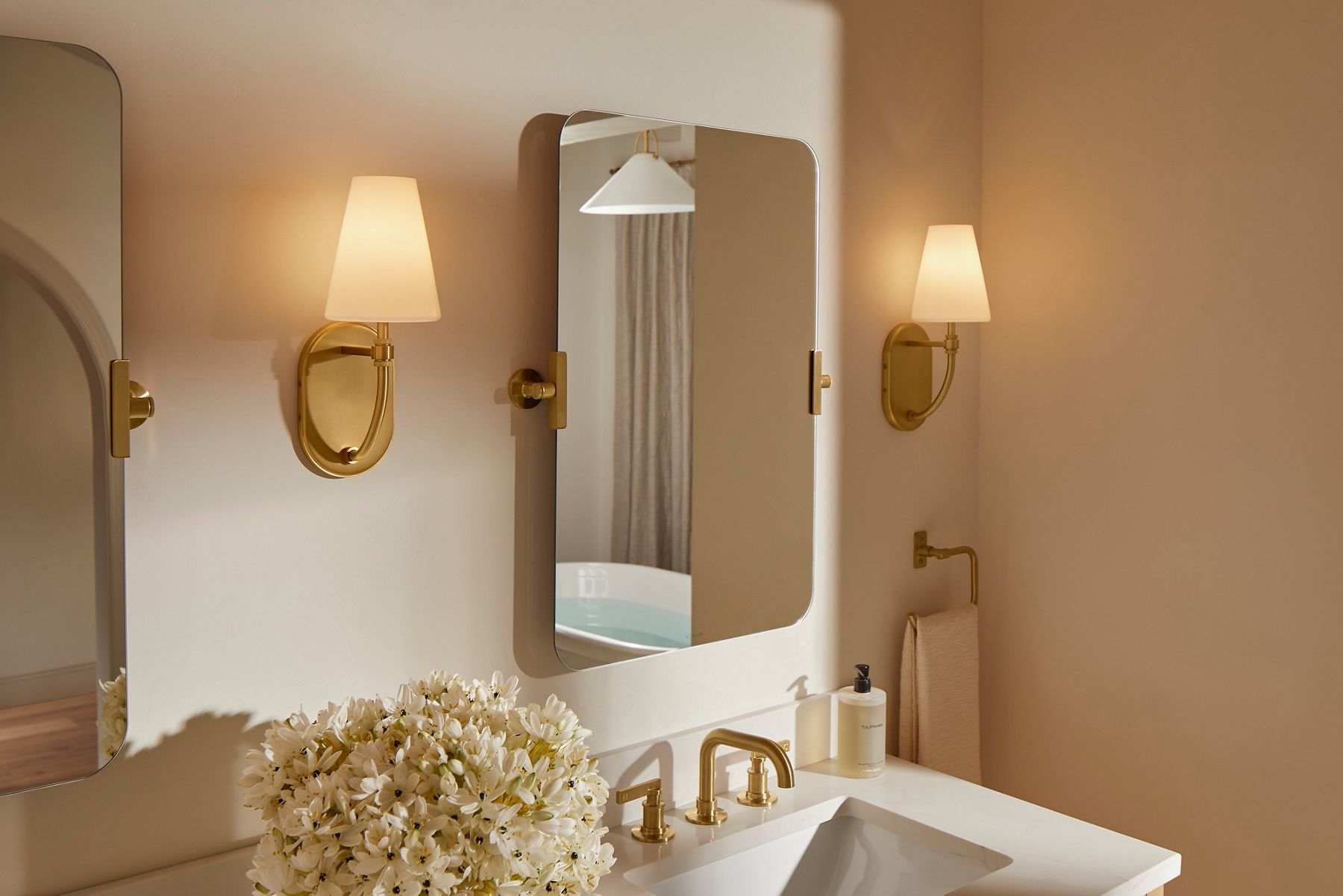 Rectangular mirrors with rounded corners and gold sconces featuring white shades above a modern sink.