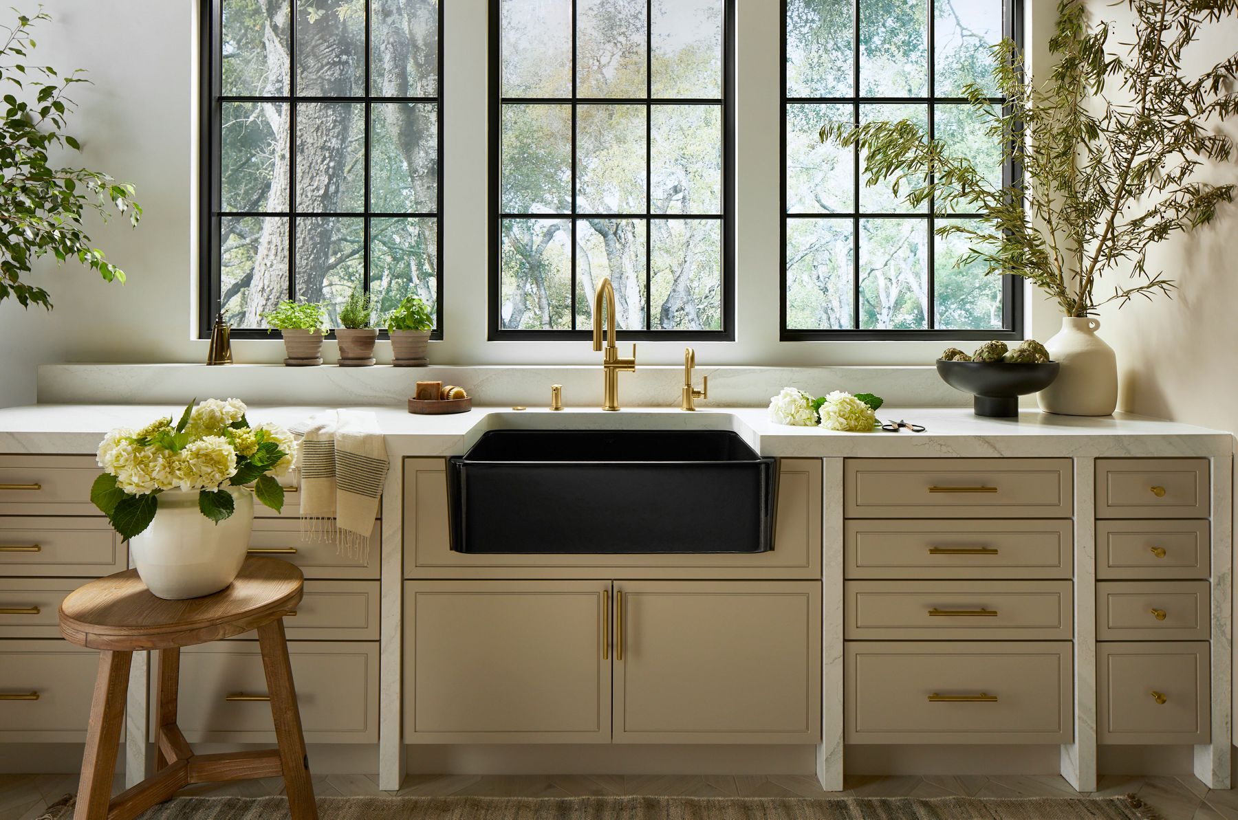 Black farmhouse sink with single-handle faucet, soap dispenser, and beverage faucet in Vibrant Brushed Moderne Brass with neutral cabinetry and countertop with windows in background.