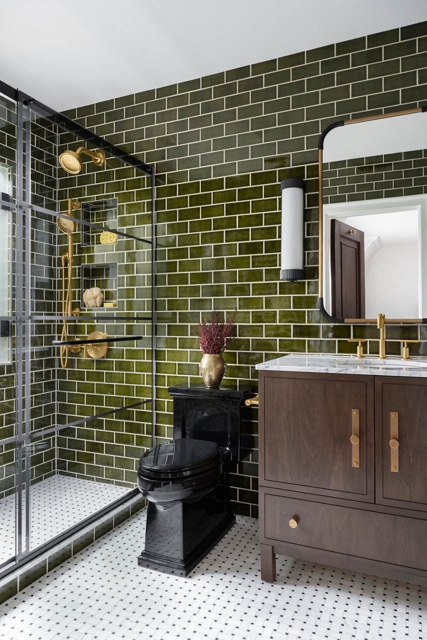A modern bathroom design featuring rich green tiled walls, a large mirror, and gold fixtures. The bathroom includes a marble countertop with dark wood cabinets and a vase with decorative branches.