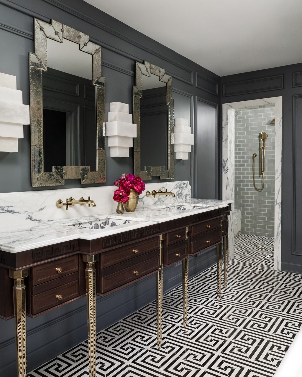 A luxurious primary bathroom featuring a marble countertop with dual sinks and gold fixtures, dark wooden cabinetry with intricate details, a vase of vibrant pink flowers, and a sophisticated timeless style reflected in the elegant decor and design.