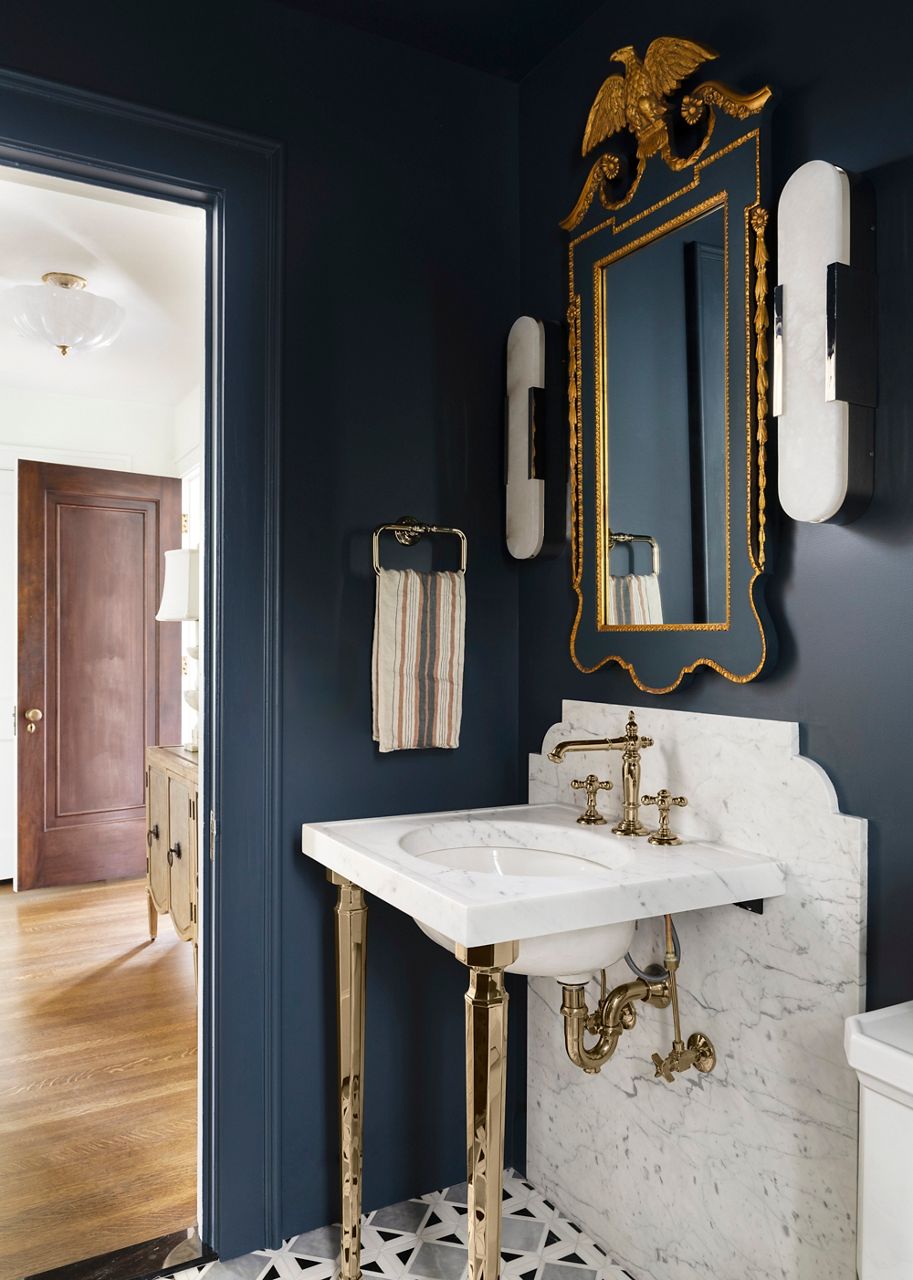 An elegant guest bathroom featuring deep blue walls, a white marble countertop with polished metal fixtures, a decorative vase with white hydrangeas, and a gold-framed octagonal mirror.