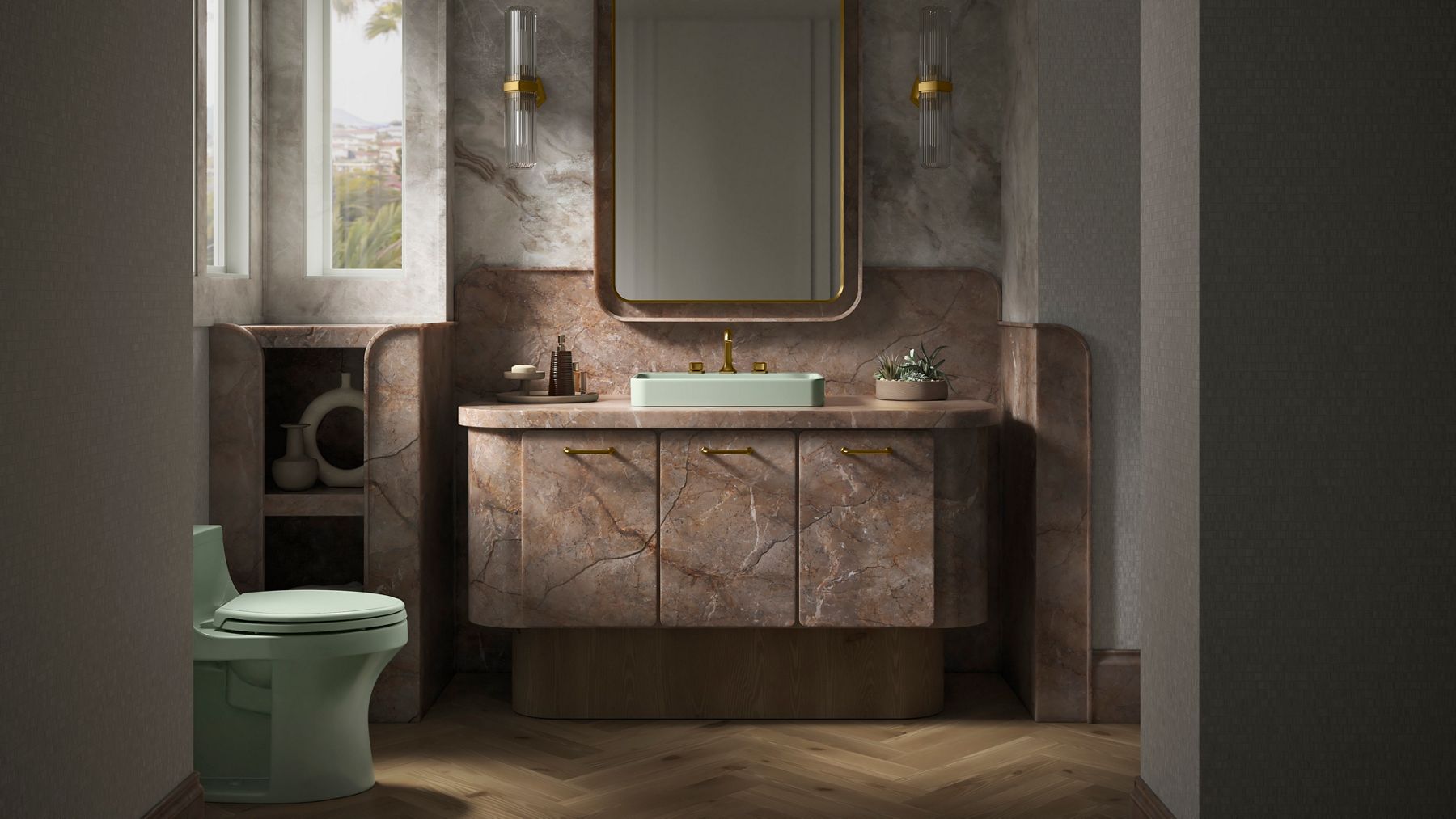 A wide shot landscape image of a sunlit bathroom designed modernly designed with rosey brown marble, beige tiled walls, oak wood flooring, and a pale green sink and pale green toilet. There is a cabinet next to the vanity with decor and towels.