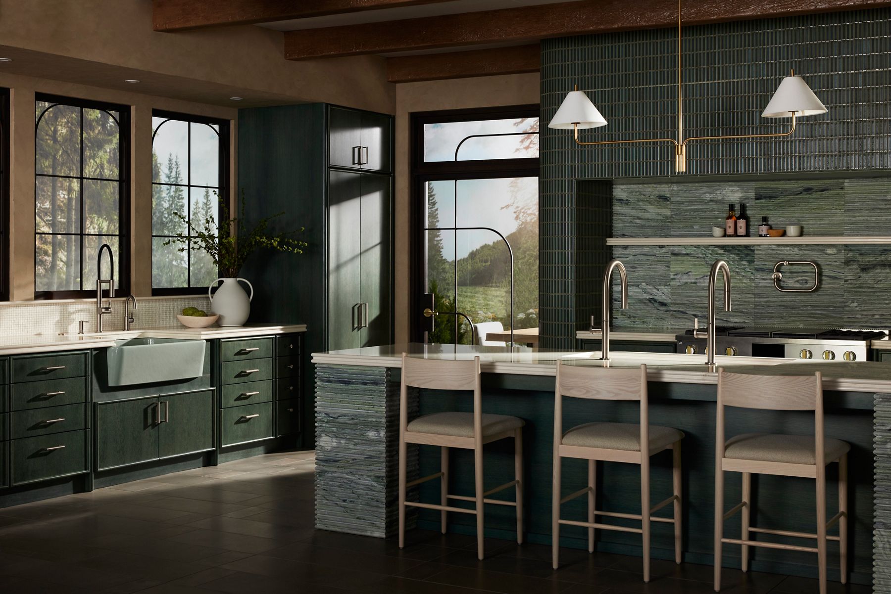 A sun drenched kitchen designed in a nature-inspired green palette. There are giant windows with greenery in the background. There is a kitchen island to the right side of the photo with an oven and kitchen counter behind it and an overhead shelf with spices neatly placed on it. The kitchen island has a double sink with silver faucets. On the left side is another kitchen space area with a farmhouse sink attached to a green kitchen counter. The sink is a pale green and the faucet is silver.