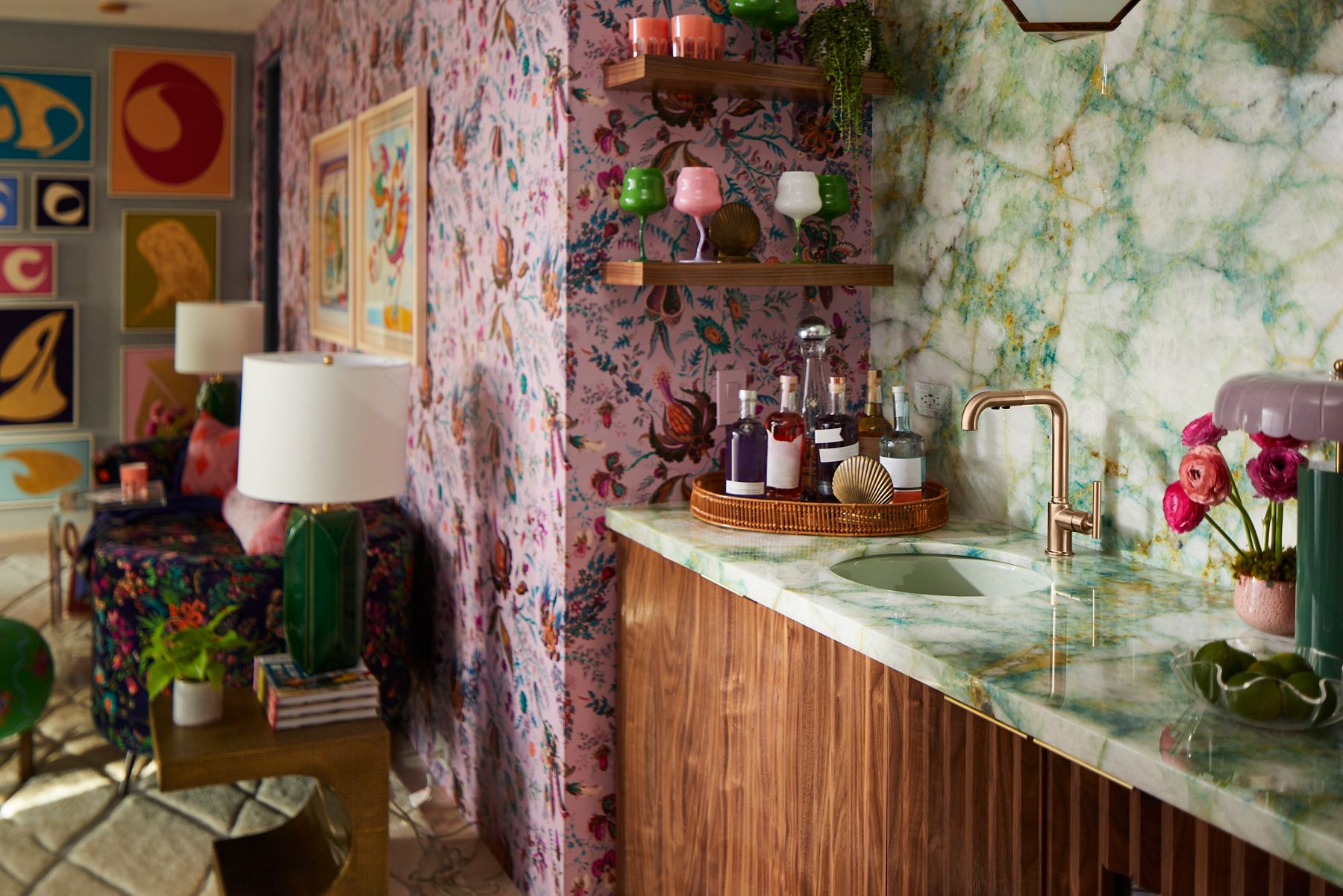 A vibrant parlour area featuring a countertop with a marble-like finish, a modern gold faucet, and a round tray holding various bottles. The background includes colorful floral wallpaper and framed artwork.