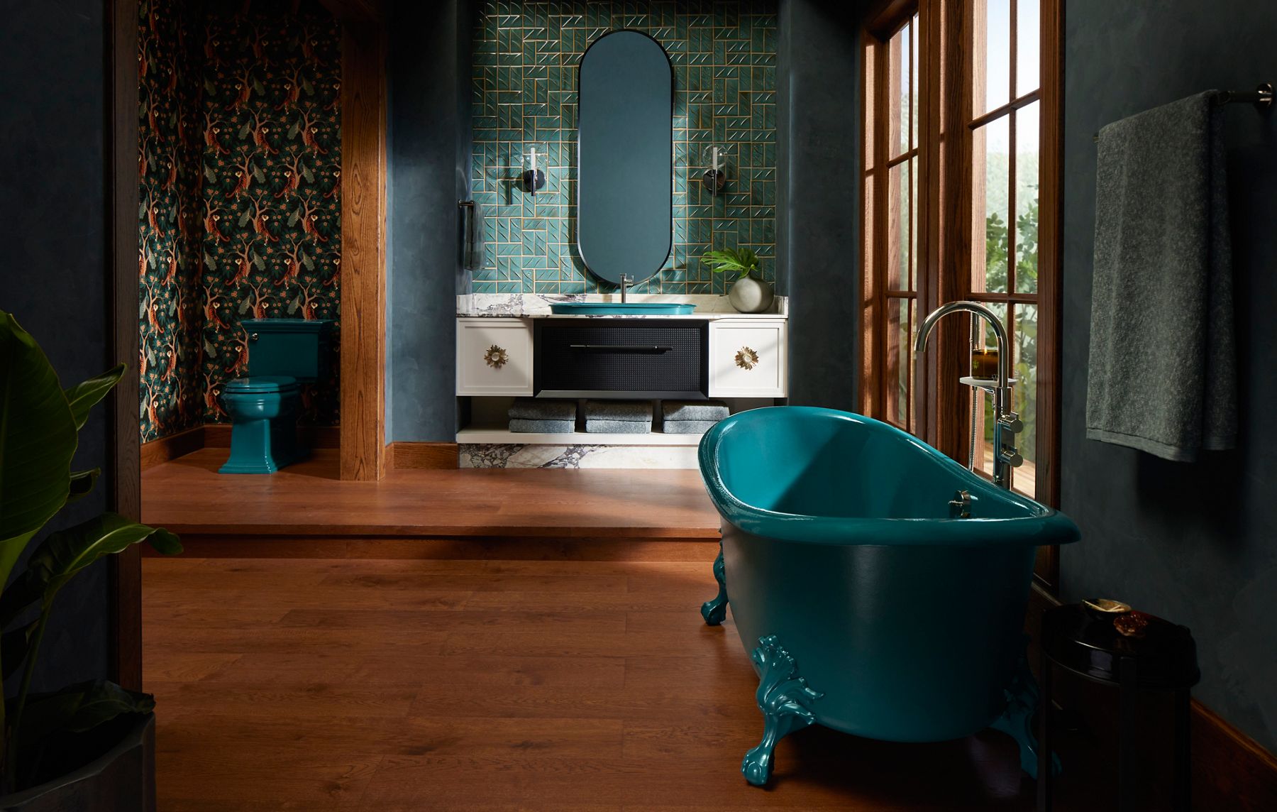 A wide shot sun drenched bathroom with a giant window to the right side of the room. There is a teal claw bath tub at the far right corner of the photo with a silver faucet. Across the tub in the back side of the room is a beautiful vanity area with a two-tone black and white vanity with gold decor detailing. Attached to the vanity is a teal sink with a silver faucet and oval mirror overhead. To the far left is a teal toilet with the lid down in an enclosed space designed in a teal tropical wallpaper.