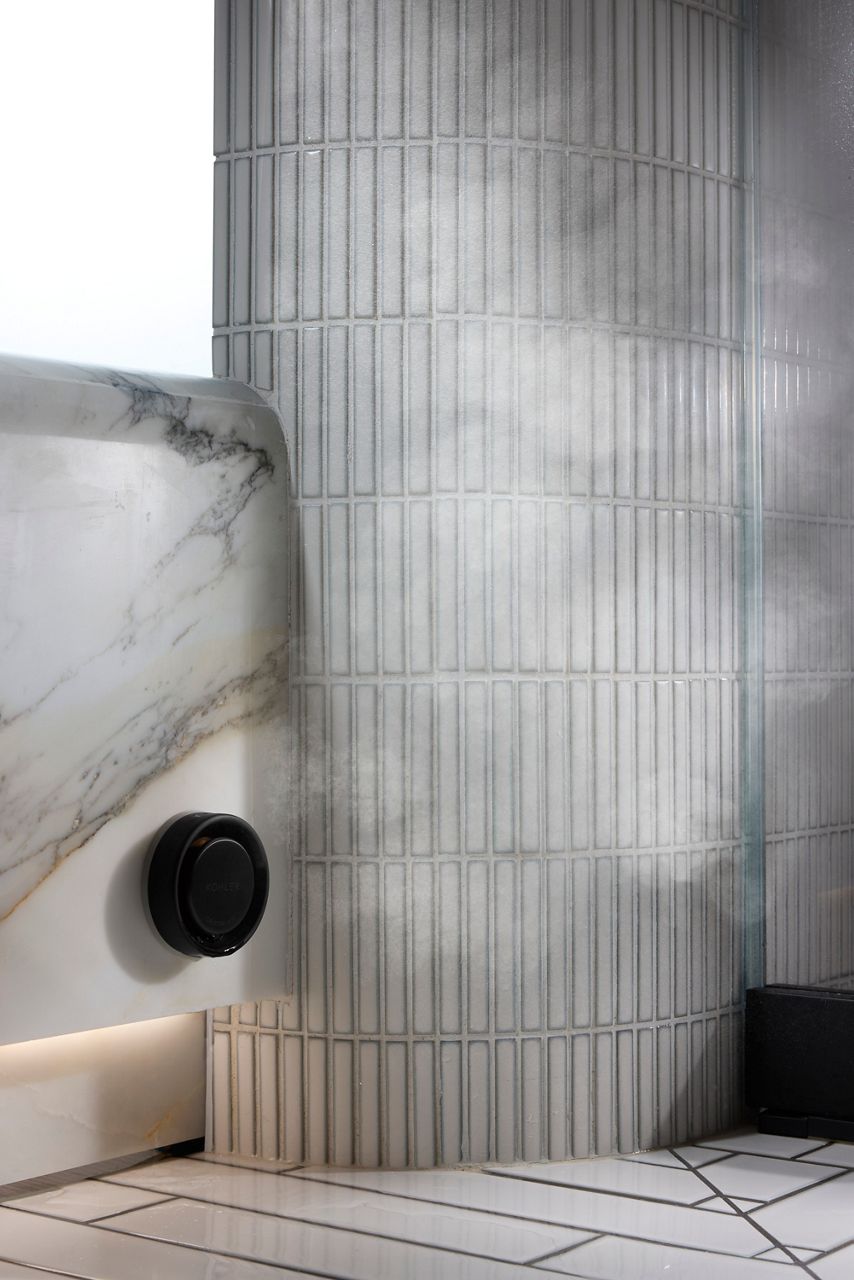 KOHLER black temperature control knob mounted on a sleek marble and tiled shower wall, with steam gently rising in the background.