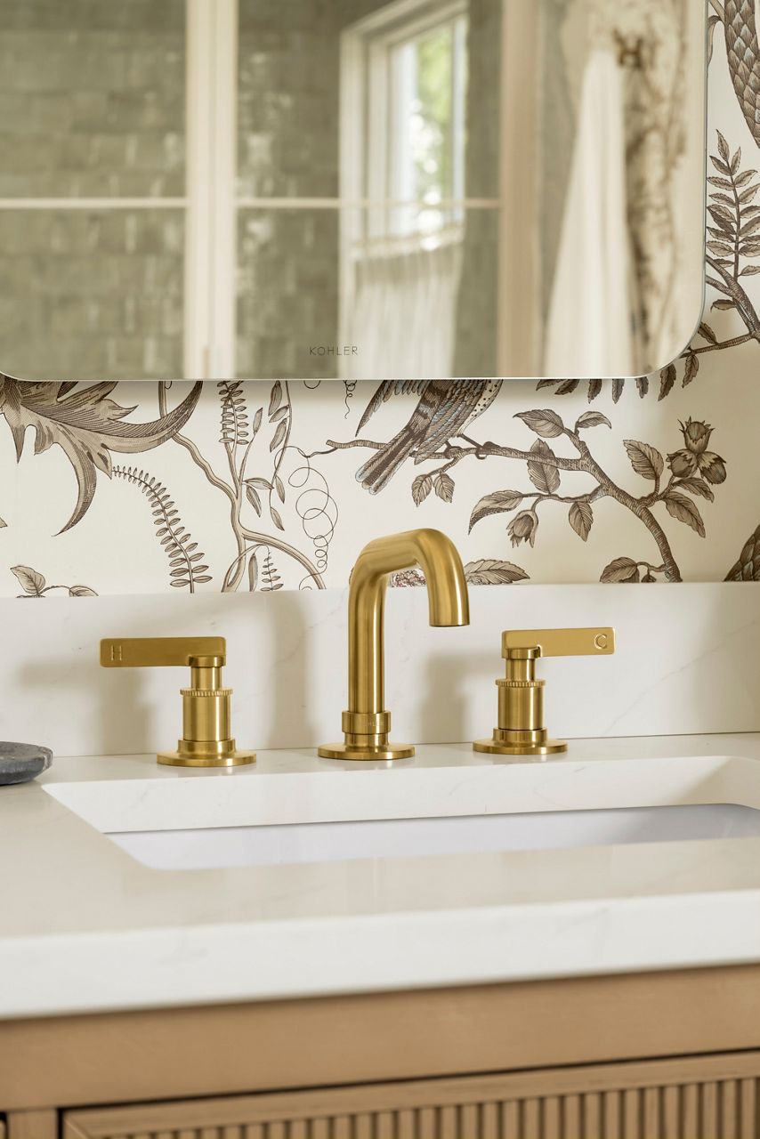 A widespread bathroom sink faucet with brushed brass finish on a white counter with wall paper and mirror in background.