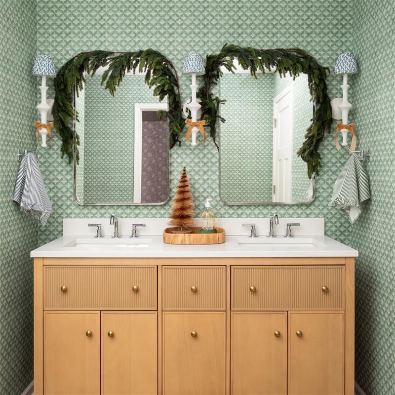 Double vanity with light wood cabinetry, white countertop, and two mirrors set against green patterned wallpaper, decorated with garland and sconces.