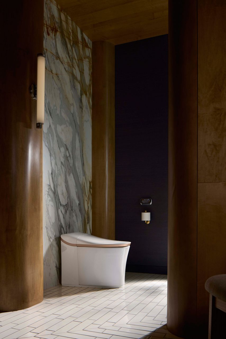 White smart toilet with Modern Brushed Brass trim with marble pattern wall, white tile floor, and wall-mounted toilet tissue holder and remote controler