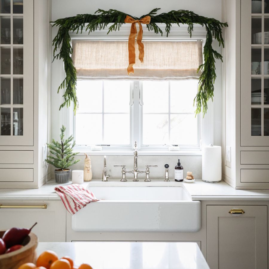 Bright kitchen with a white farmhouse sink beneath a window, framed by cabinetry and greenery garland with an orange ribbon above.