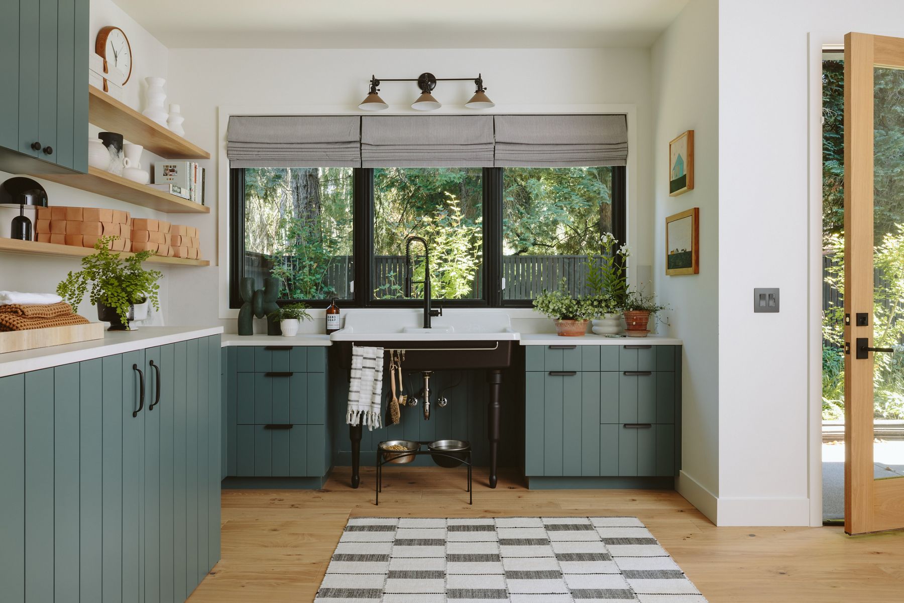 Cozy kitchen with muted green cabinetry, open wood shelves, and a large window overlooking greenery.