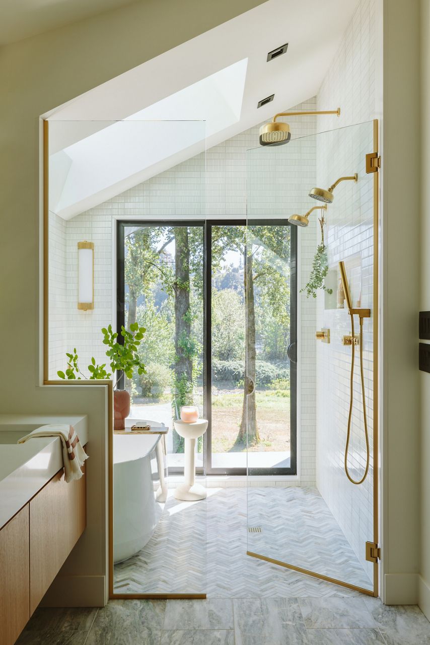 Spacious contemporary bathroom with a glass-enclosed shower, freestanding tub, and large windows overlooking greenery, accented with warm wood and brass finishes.