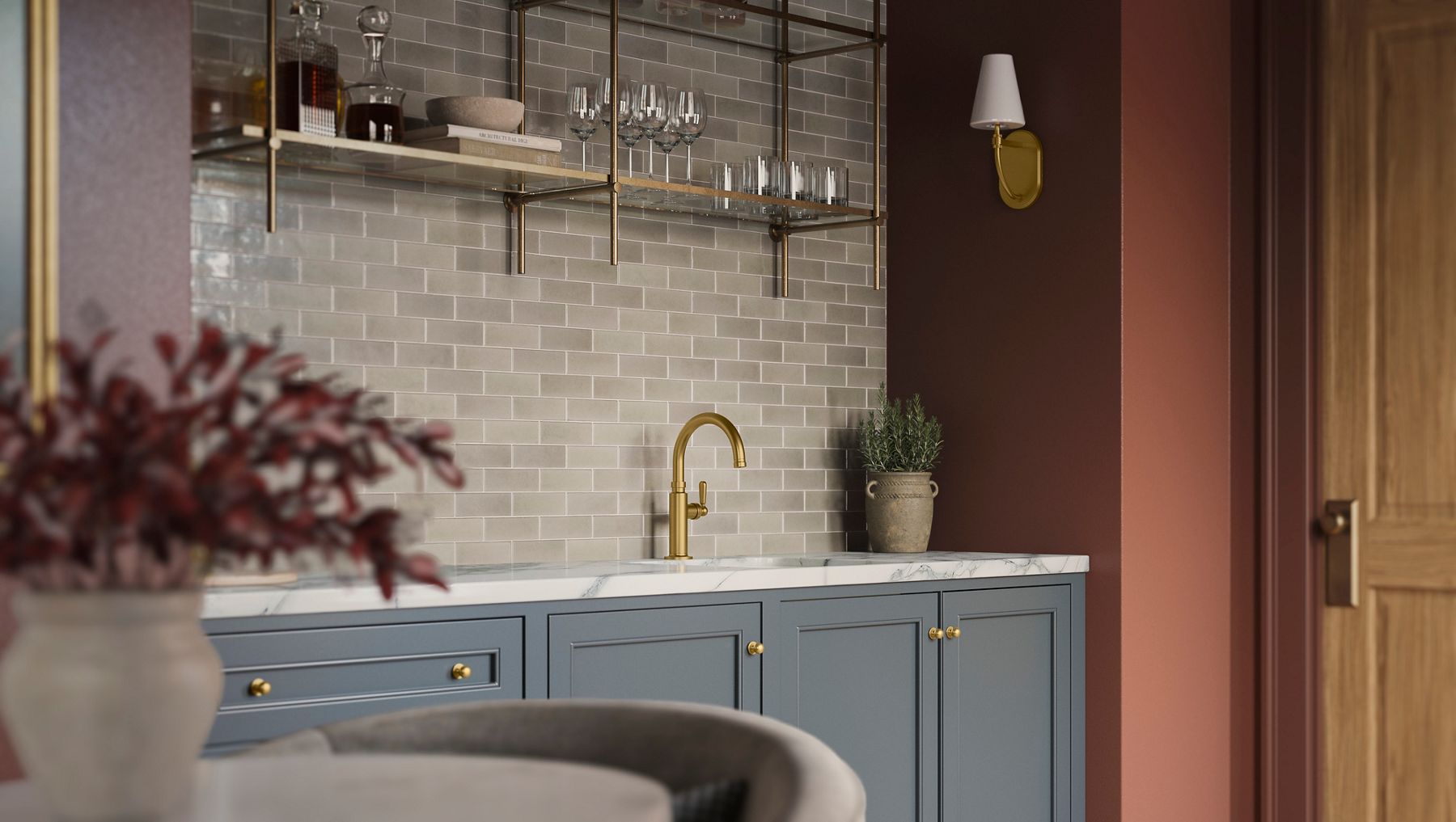Blue-gray cabinetry with brass hardware and a marble countertop, featuring a small integrated sink with a gold faucet, gray subway tile backsplash, open shelving above, and a potted plant on the counter.