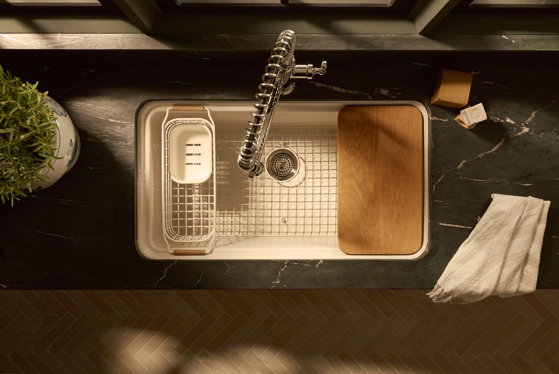 Top view of farmhouse kitchen sink with brass spring faucet, cutting board, and drying rack on dark stone countertop