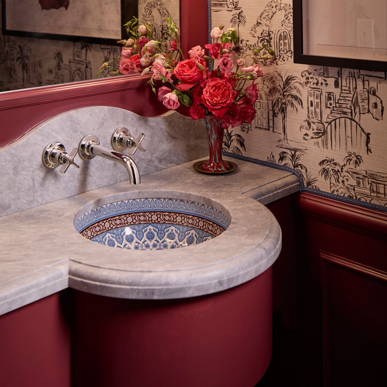 Curved red vanity with a marble top and inset patterned sink, paired with wall-mounted faucets and a vase of red flowers.
