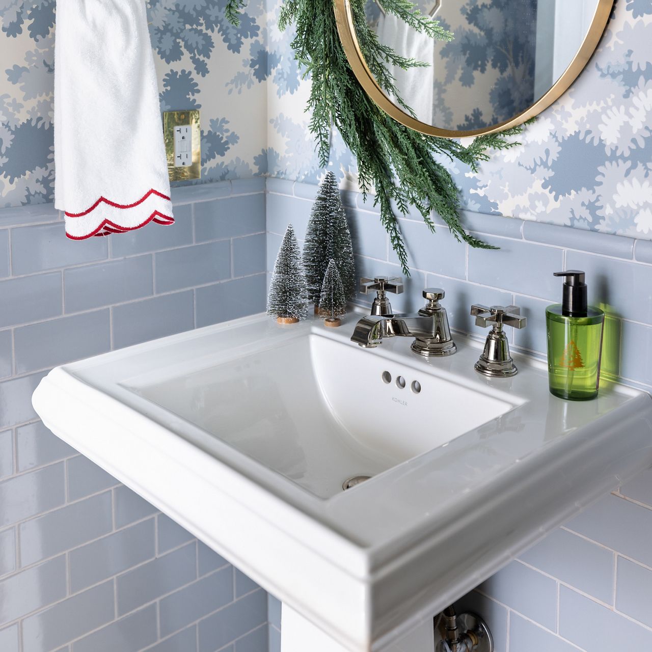 White pedestal sink with chrome faucet set against light blue tile and patterned wallpaper, decorated with greenery garland, small tabletop trees, and a hanging towel.
