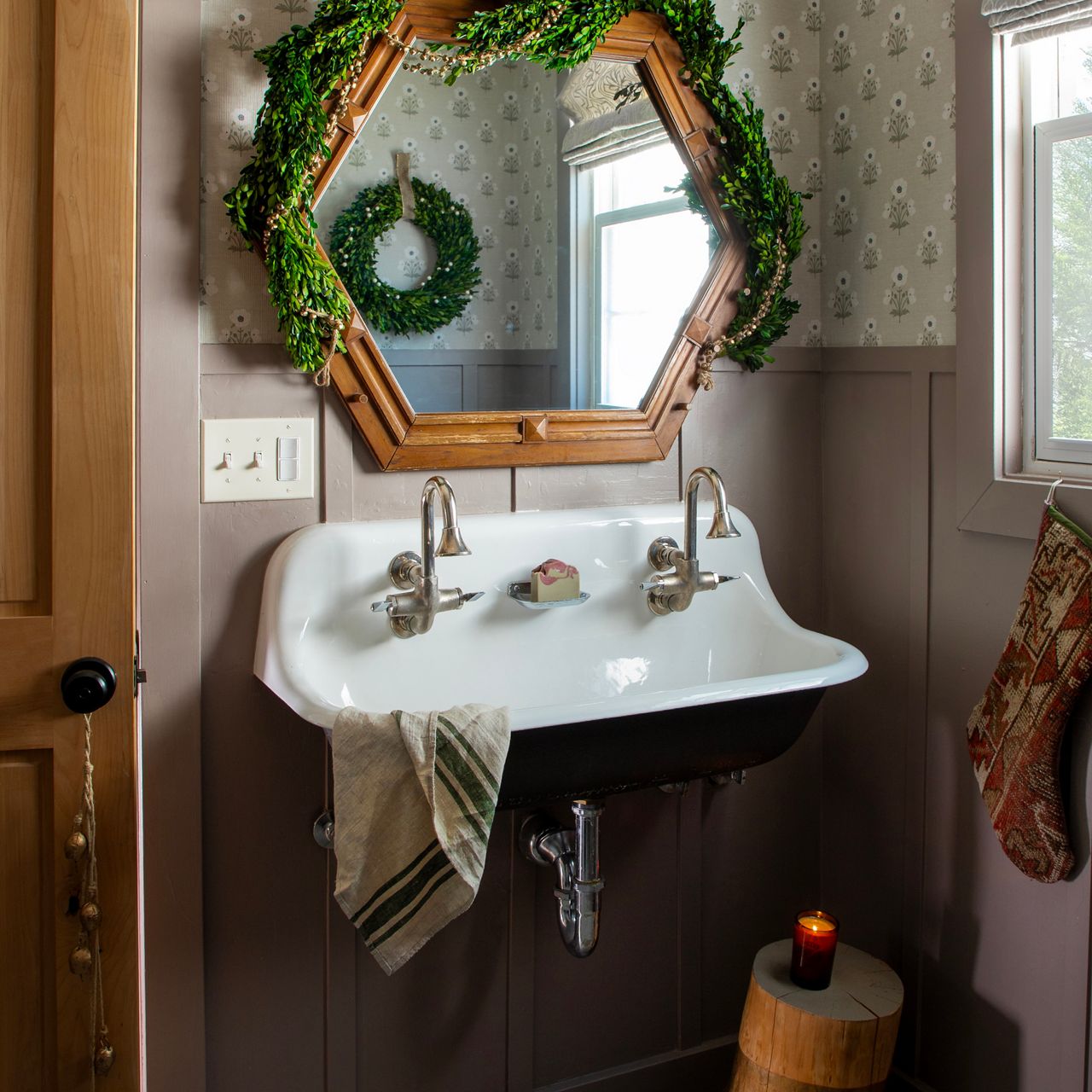 Vintage wall-mounted sink with chrome faucets beneath a geometric wood-framed mirror, decorated with green garland and a small wreath in a cozy, softly lit bathroom.