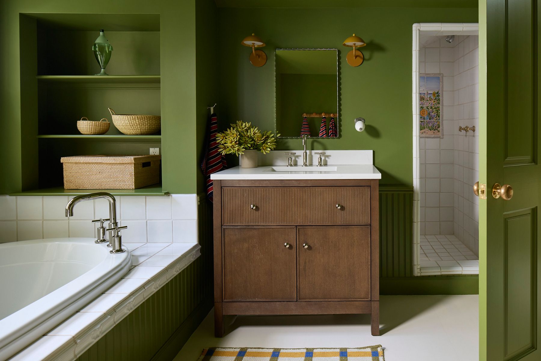 Moody green bathroom featuring a dark wood vanity with white sink, built-in shelving, brass sconces, and a bathtub in the foreground.
