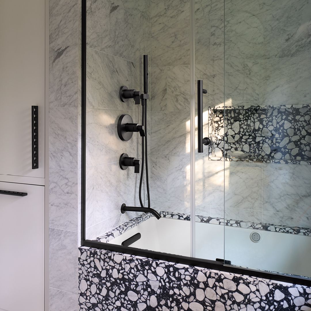 Bathtub-shower combination with glass doors, black fixtures, marble-look walls, and a terrazzo-style tub surround.