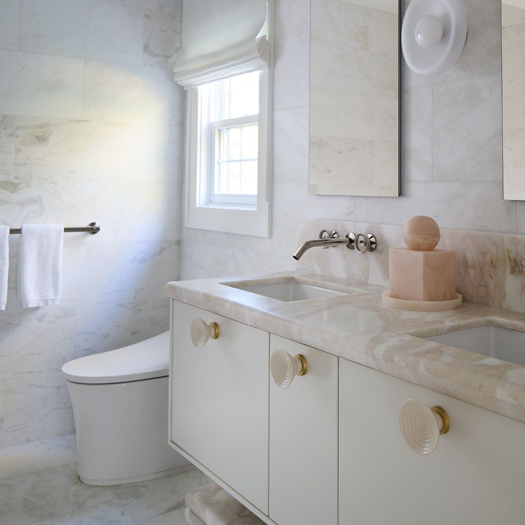 Bright marble bathroom featuring a double vanity with light cabinetry, round knobs, wall-mounted faucets, and a nearby toilet under a sunlit window.