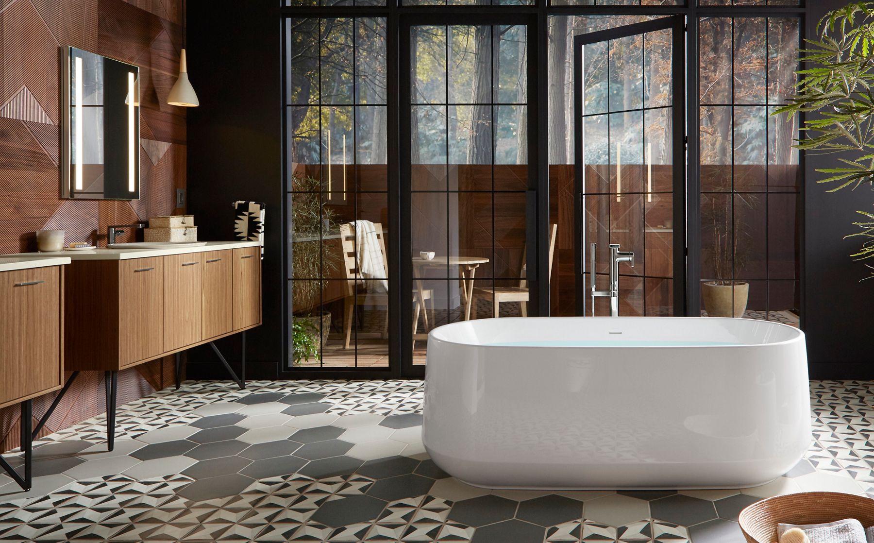 A white KOHLER Ceric bath stands in the middle of a bathroom with wood paneled wall, two wall mounted Jute vanities, patterned tile floors, and floor to ceiling windows overlooking a balcony.