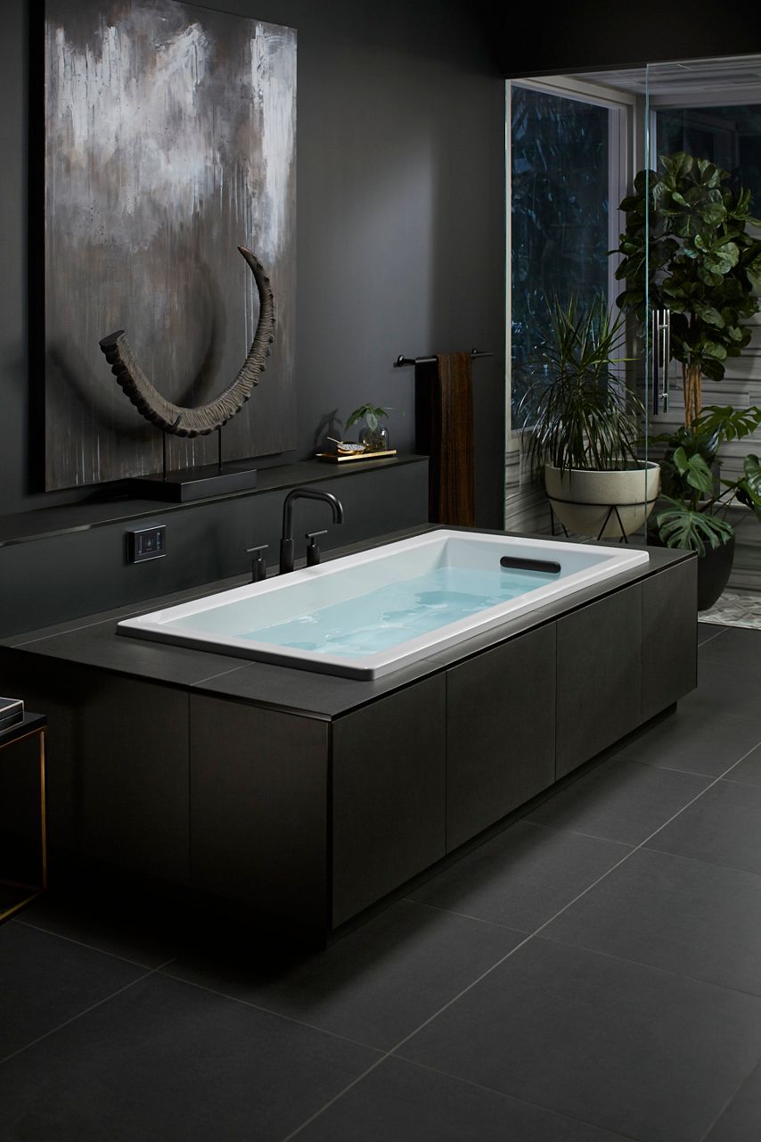 Image of an elongated rectangular white KOHLER sink installed in a dark black themed bathroom design. It's filled with water and indicates its ability to offer sensory experiences with water in the sink.