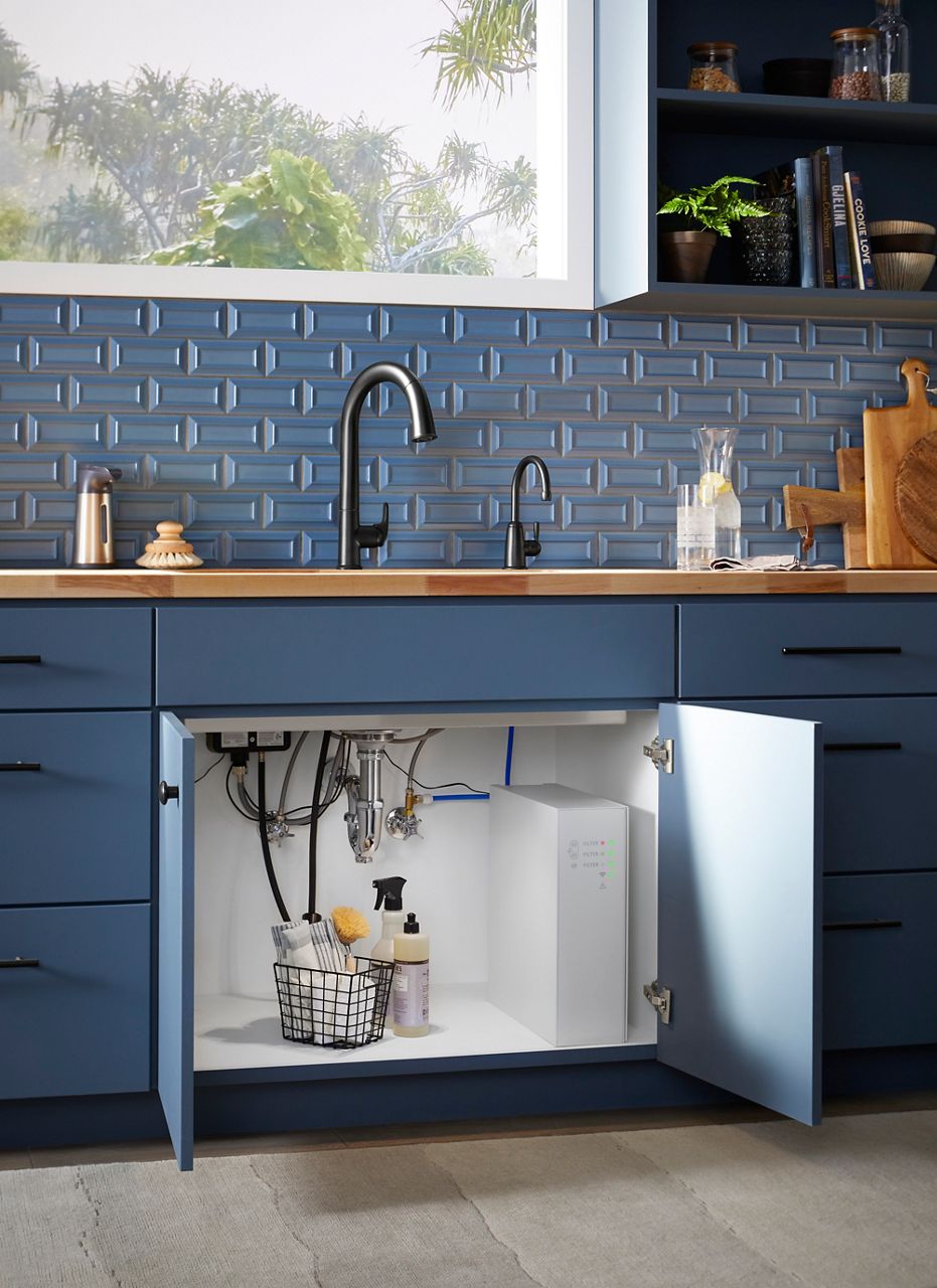 A blue kitchen cabinet with doors open under the sink, showing a white box with green lights that is a water filter.