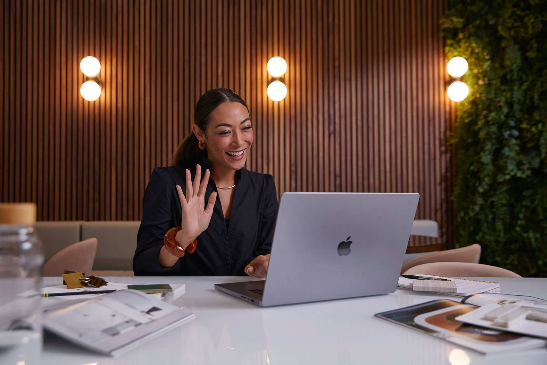 A smiling woman in a video call on a laptop, seated at a desk with design materials in a room with wooden paneling and the overlay text “KOHLER Bathroom Design”.