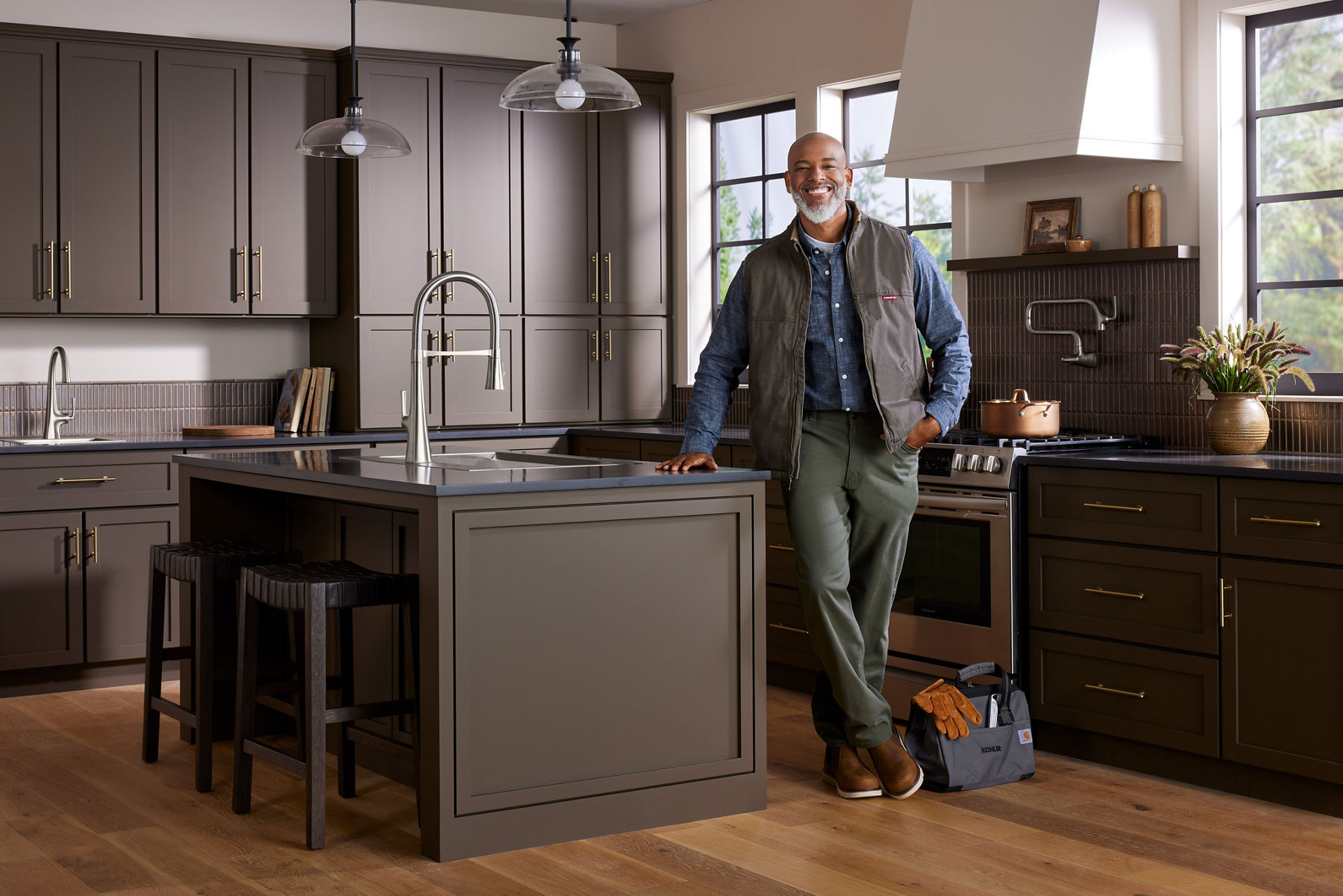 A smiling man stands casually in a modern kitchen next to a large island, promoting installation services. He wears a vest over a shirt with olive-green pants and brown shoes. The kitchen features sleek gray cabinets, pendant lighting, and modern appliances.