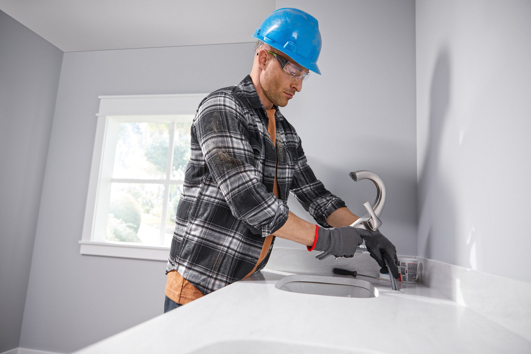 A man in a hardhat and plaid shirt installs a vibrant brushed nickel bathroom faucet into a white vanity top in a pale grey bathroom.