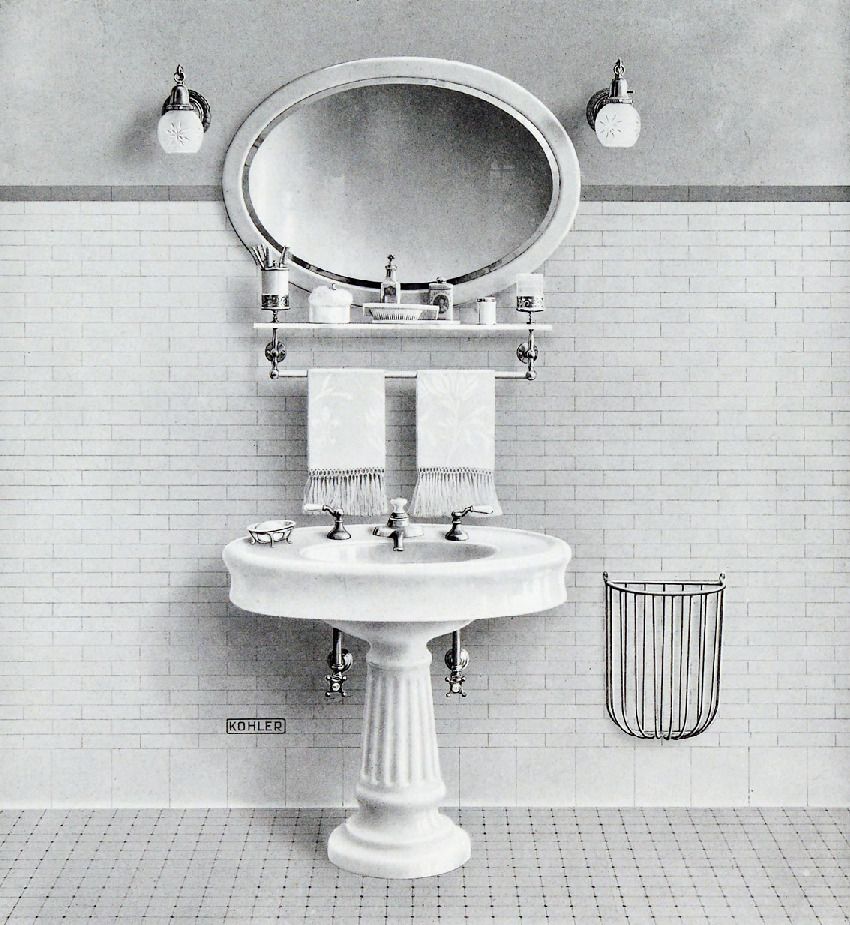 A historical black and white image of a vanity area with a pedestal sink, mirror, sconce lights, and various grooming items.