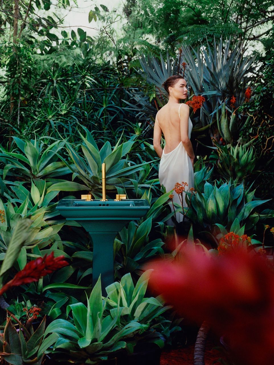 Woman wearing a white dress in a garden with teal pedestal sink with gold faucet in tropical setting