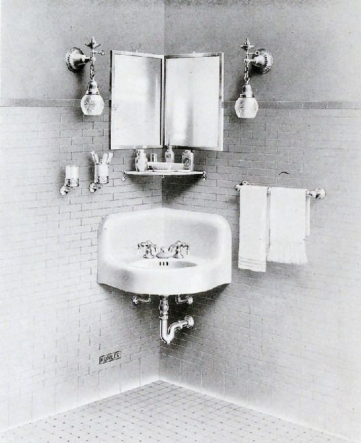 A historical black and white image of a vanity area with a corner sink and mirror, sconce lights, and various grooming items.