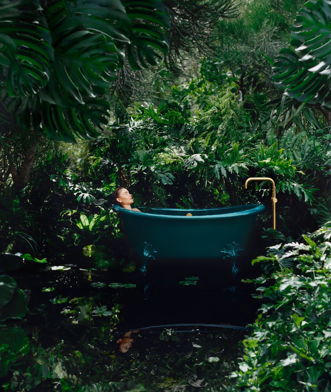 A far away shot of a woman lying inside of a teal claw tub in the middle of the forrest. The tub is positioned in the center and surrounded by greenery and water. The bath tub faucet is gold.