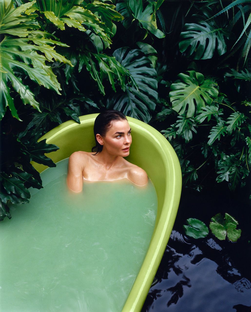Woman soaking in light green bathtub surrounded by tropical plants