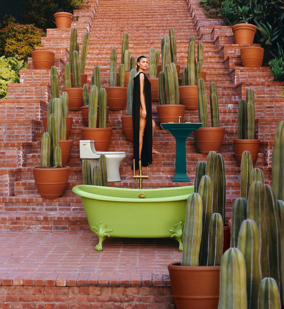 A wide shot lifestyle image taken on a long brick stairwell. A woman is standing at the center in a black dress and positioned at her side. There are potted cacti around her and a bright green claw tub at the front of the image. There's a teal kitchen sink on her right side and a pale green toilet behind her.
