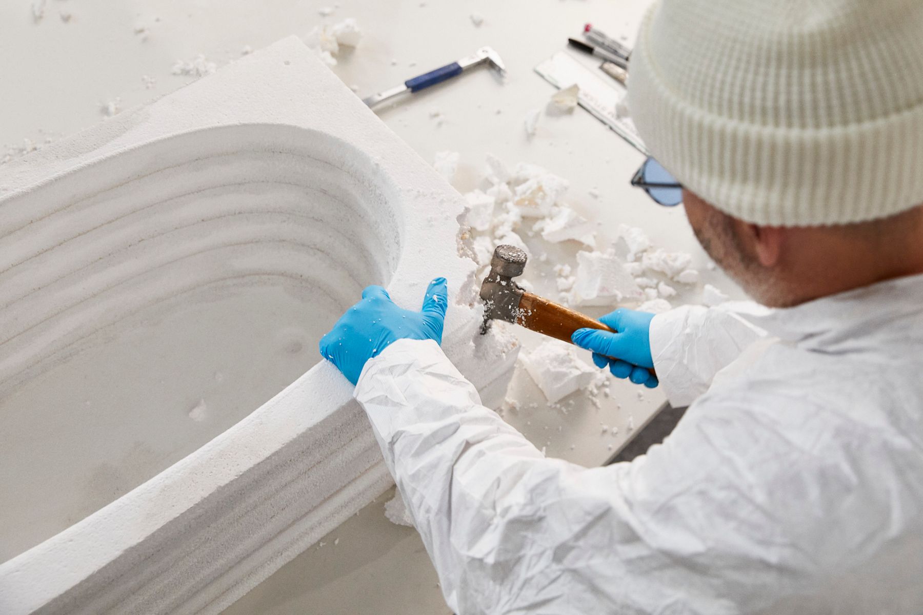 Person in protective gear, including a white suit, blue gloves, and a beanie, using a hammer to carve a piece of white foam or stone in a workshop setting. Debris and carving tools are scattered on the workbench.