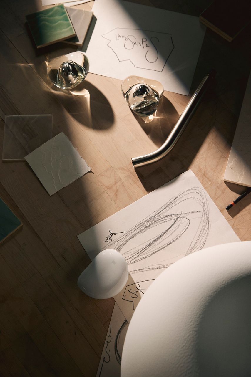 Close-up of two artistic glass knobs placed on a wooden table, surrounded by design sketches and a green ceramic tile sample. Sunlight casts shadows, highlighting the unique shapes and textures of the knobs.