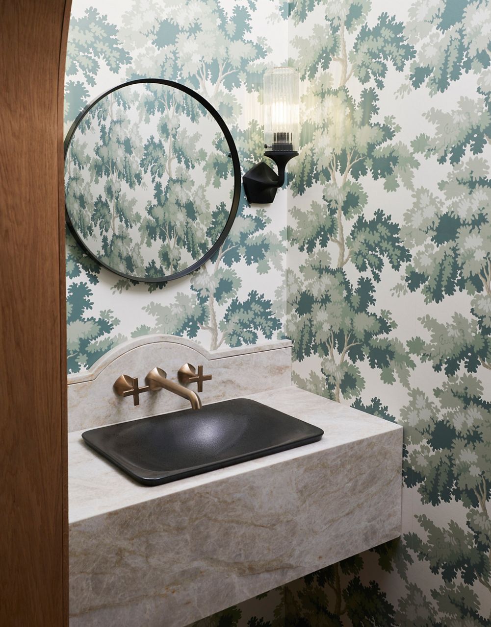 Floating stone vanity with a black vessel sink and wall-mounted brass faucet set against blue floral wallpaper and a round mirror.